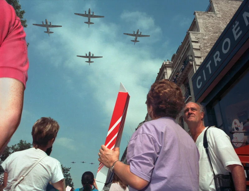 People look up at four airplanes flying in formation over a city street next to a building with a large Citroën sign.