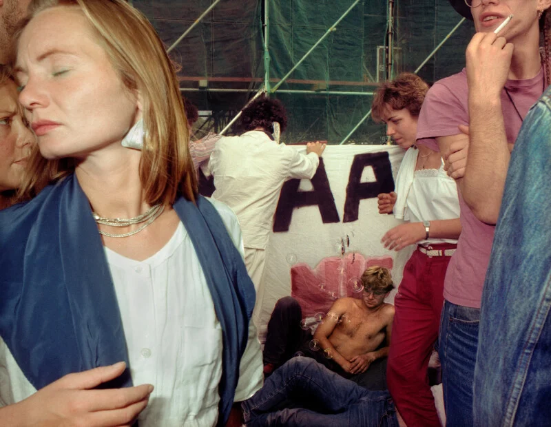 A group of people gather outdoors near a banner with the letters "AA," while a shirtless man sits on the ground nearby.