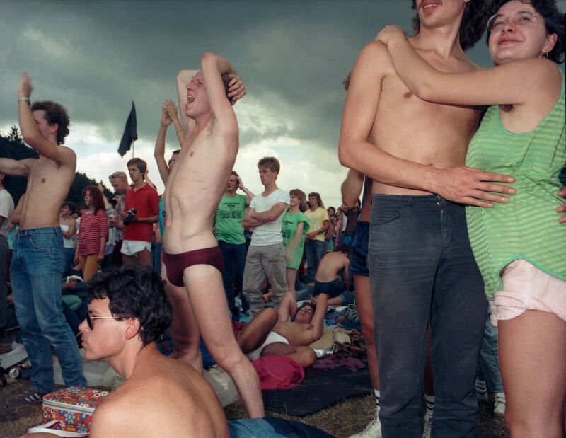 A crowd of young people at an outdoor festival, with some standing and others sitting on the grass under a cloudy sky.