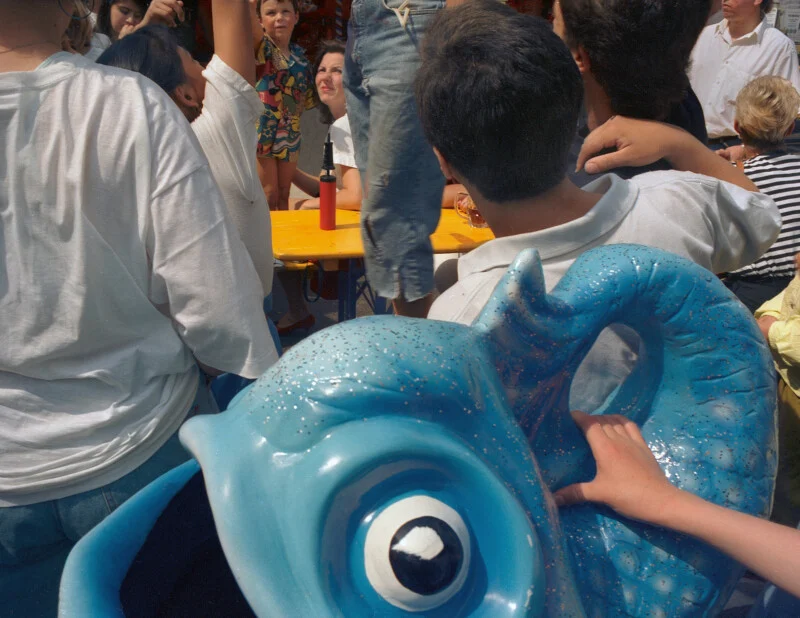 A close-up view of a large, glittery blue seahorse-shaped ride seat in the foreground, with a crowd of people standing and sitting around a yellow table in the background.