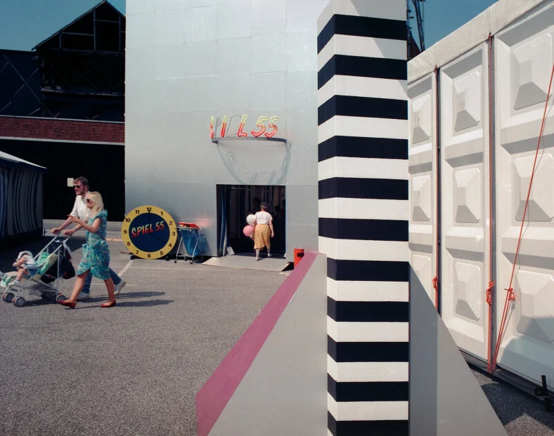 A family walks past a silver building marked "SPIEL 55" with a large clock-shaped sign, next to a black-and-white striped pillar.