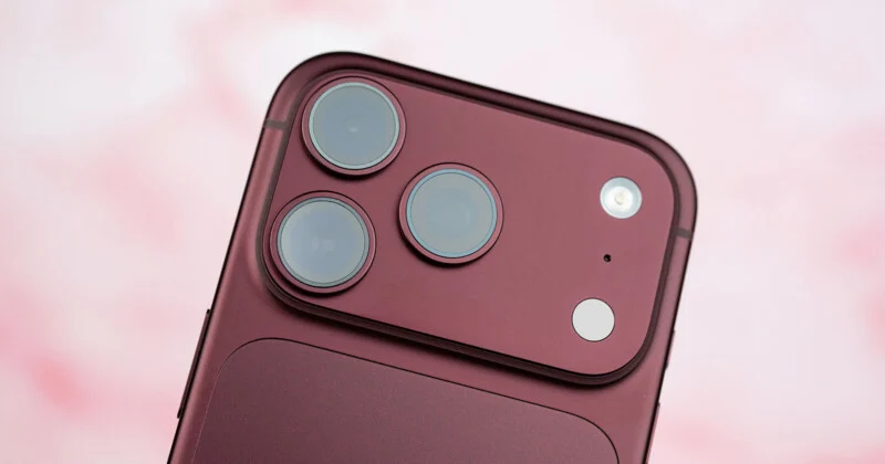 A close-up view of the back of a deep red smartphone, showing a raised camera module with three circular lenses, a flash, and a small sensor against a soft pink background.