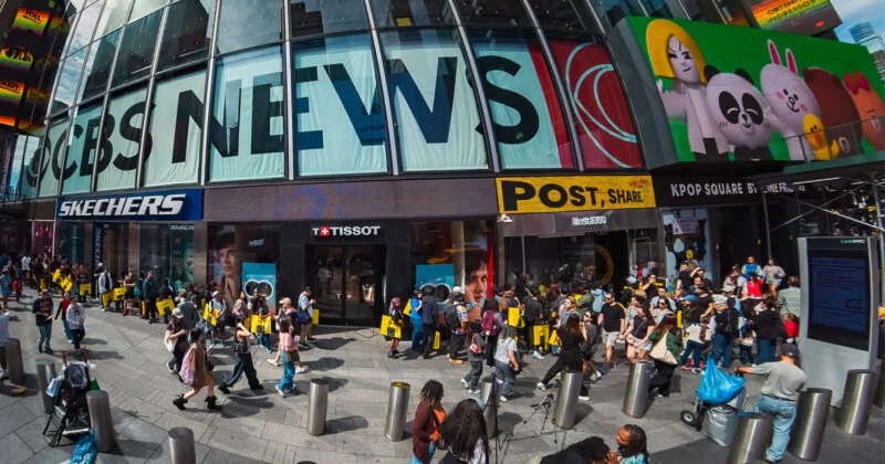 A wide-angle view of a busy city street with pedestrians walking past storefronts featuring large signs for CBS News, Skechers, Tissot, and Line Friends.