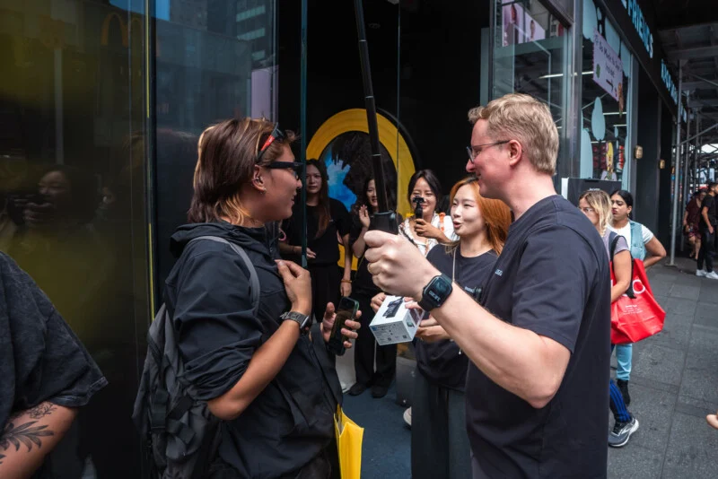 A man holding a selfie stick and a product box speaks to a person holding a smartphone outside a storefront, with several other people standing nearby.