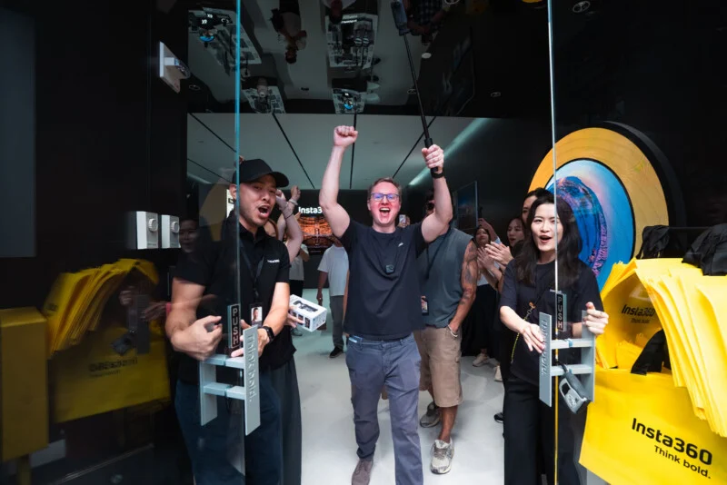 A group of people cheer as they walk through glass doors into an Insta360 store, with one man in the center holding a camera on a selfie stick.