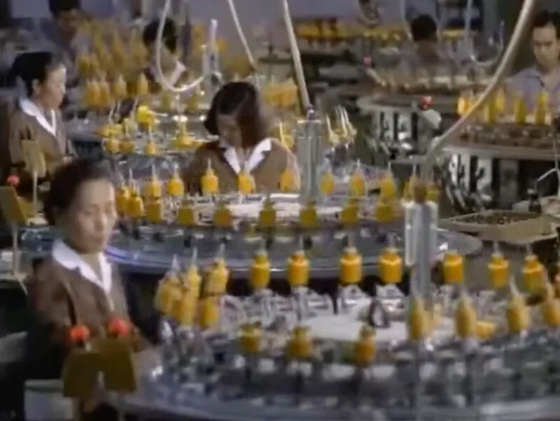 Several workers in a factory operate circular machinery with many small, yellow, upright components arranged in a ring.
