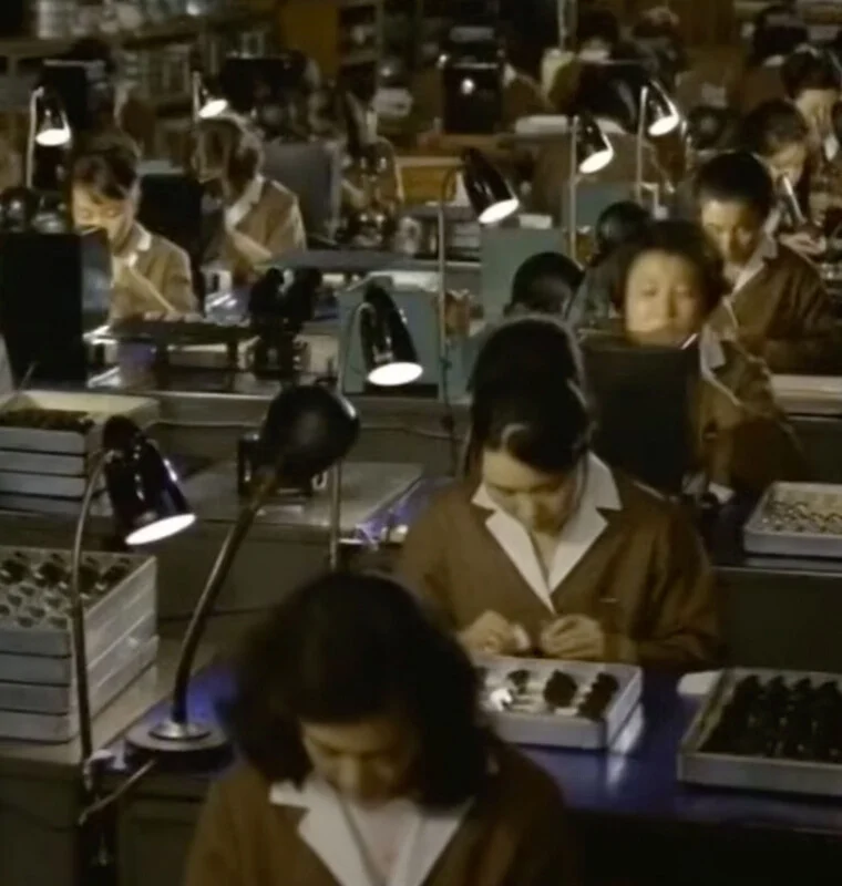 A high-angle shot shows rows of women in brown uniforms working at desks with individual lamps, assembling small components in trays.