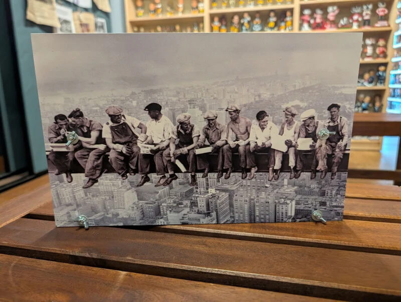 A black-and-white print of the iconic "Lunch atop a Skyscraper" photograph mounted on a wooden stand with four metal wing nuts.
