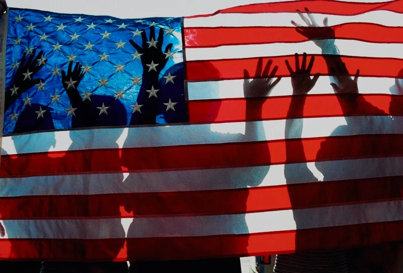 Silhouettes of people with their hands raised are visible behind a large, translucent American flag.
