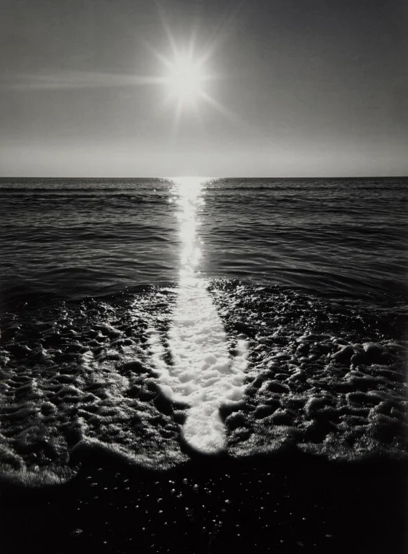 A bright sun shines above the horizon, casting a vertical reflection across the dark ocean water onto the foamy shoreline.