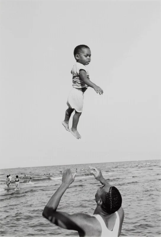 A man in a white tank top tosses a young child into the air above the ocean, with other people swimming in the background.