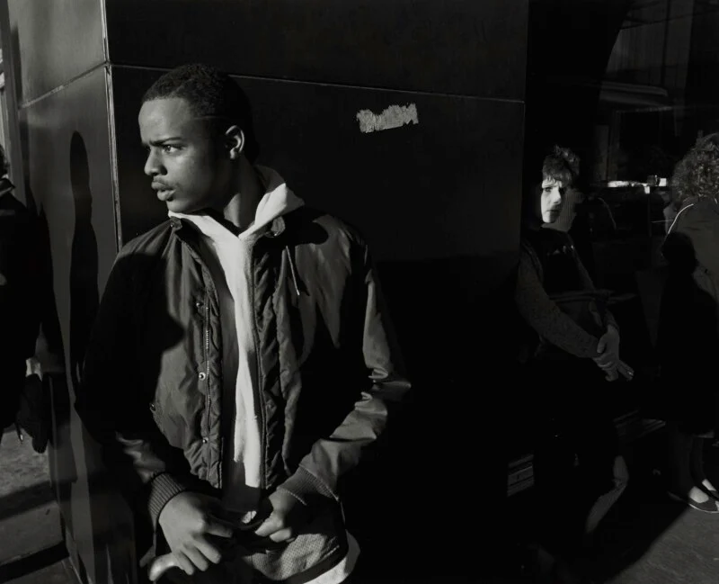 A young man in a jacket and hoodie stands in the foreground, looking to the left, while a person stands in the shadows behind him.