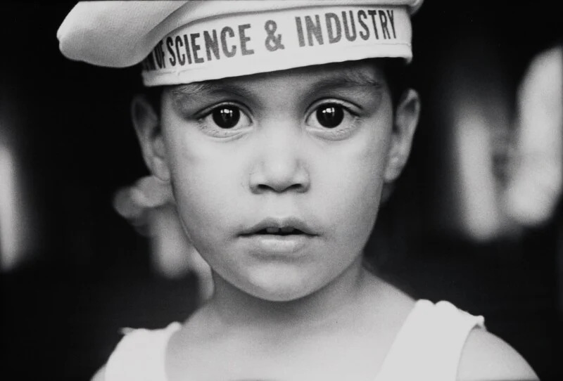 A close-up, black-and-white portrait of a young child wearing a white sailor-style cap labeled "SCIENCE & INDUSTRY.