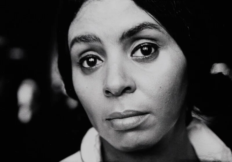A close-up, black-and-white portrait of a woman with dark hair looking directly at the camera with a neutral expression.