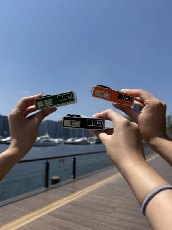 Three hands hold up small, rectangular green, orange, and black cameras against a background of a harbor and blue sky.