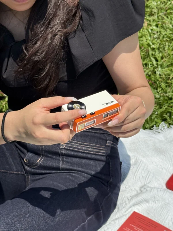 A person with manicured nails holds a small, rectangular white and orange Yashica Y16 camera while sitting on a blanket outdoors.
