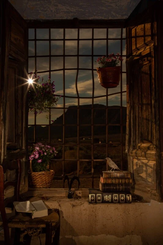 An open wooden window with iron bars overlooks a mountain landscape at sunset, featuring a chair with an open book, potted flowers, and a stack of books topped with a bird figurine.