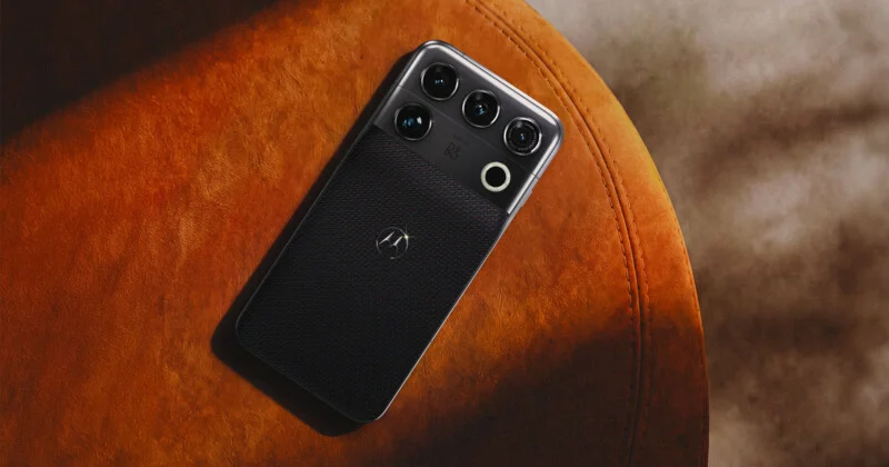 A black smartphone with a textured back and a prominent camera module featuring three lenses and a circular flash, resting on an orange surface.