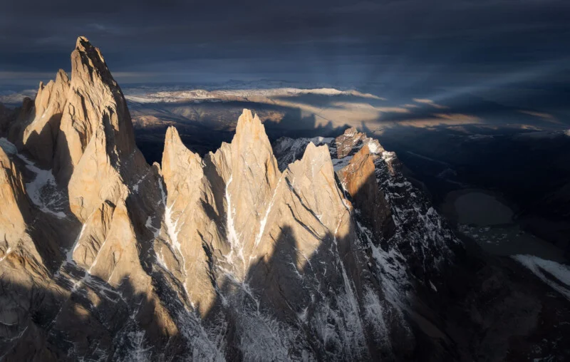 Golden sunlight illuminates the sharp, snow-dusted granite peaks of a mountain range against a dark, dramatic sky.