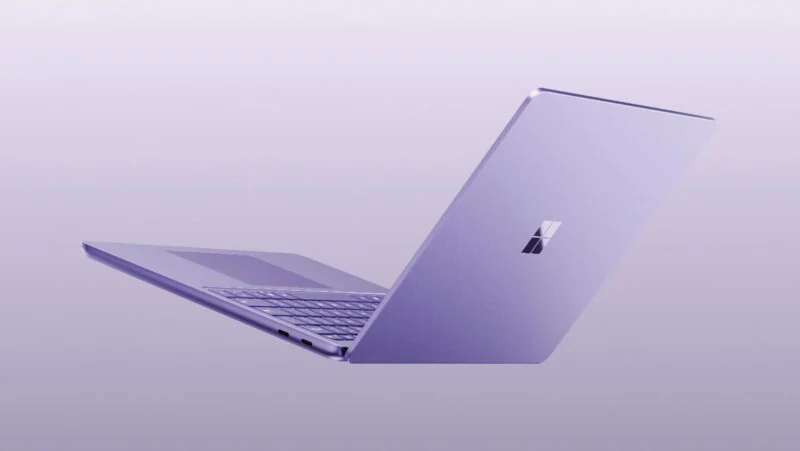 A light purple laptop is angled open against a matching light purple background, showing the back of the lid with a Microsoft logo and a partial view of the keyboard and trackpad.
