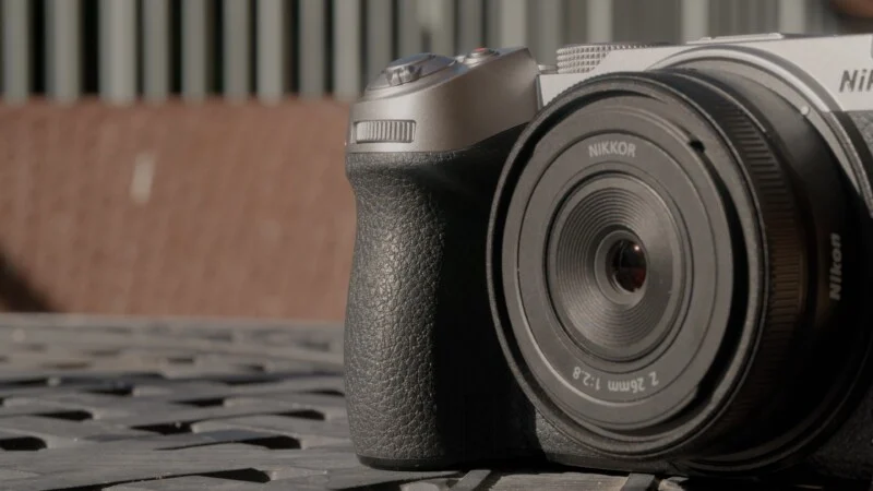 A silver and black Nikon mirrorless camera with a NIKKOR Z 26mm f/2.8 lens rests on a dark, textured surface.