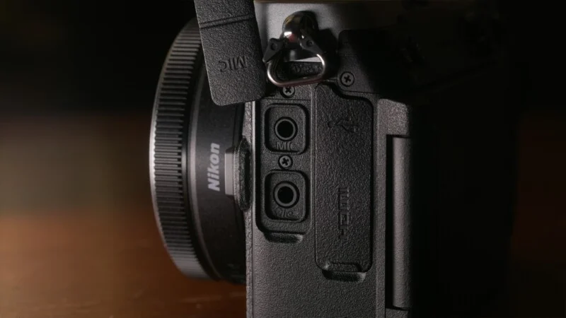 A close-up view of the side of a Nikon camera, showing open rubber port covers for microphone and headphone jacks, with a larger closed port cover labeled with USB and HDMI icons.