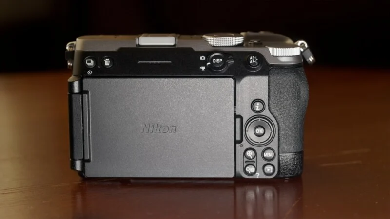 The back of a black Nikon mirrorless camera sits on a wooden surface, showing its LCD screen, control buttons, and dials.