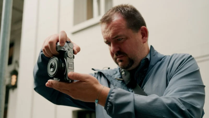Een man met een sikje en een blauwe jas houdt een zilveren digitale camera stevig vast terwijl hij naar het scherm kijkt.