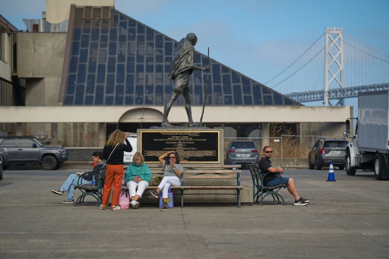 A bronze statue of Mahatma Gandhi stands on a pedestal behind a park bench where four people are sitting and one is standing, with the San Francisco-Oakland Bay Bridge visible in the background.