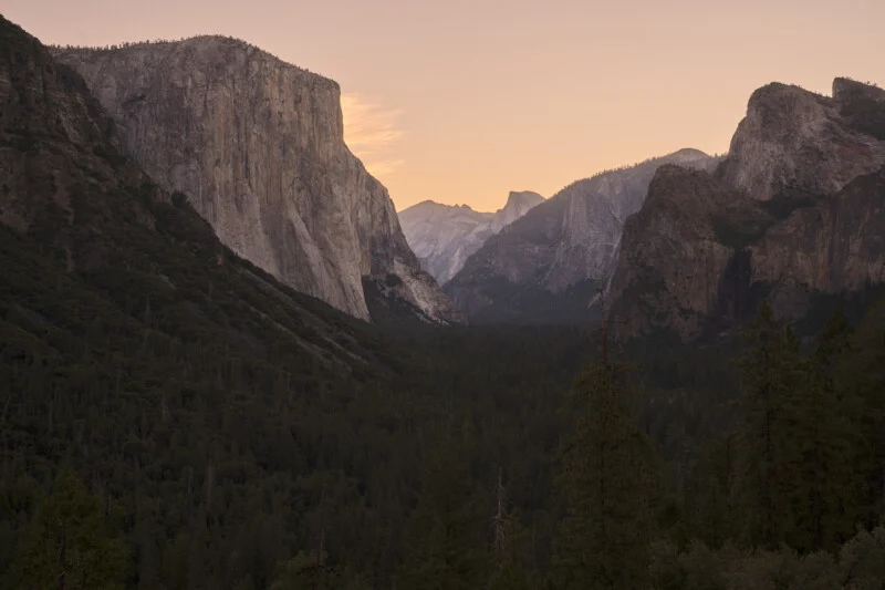 A vast valley filled with dense forest is framed by towering, steep granite cliffs under a pale, sunset-lit sky.