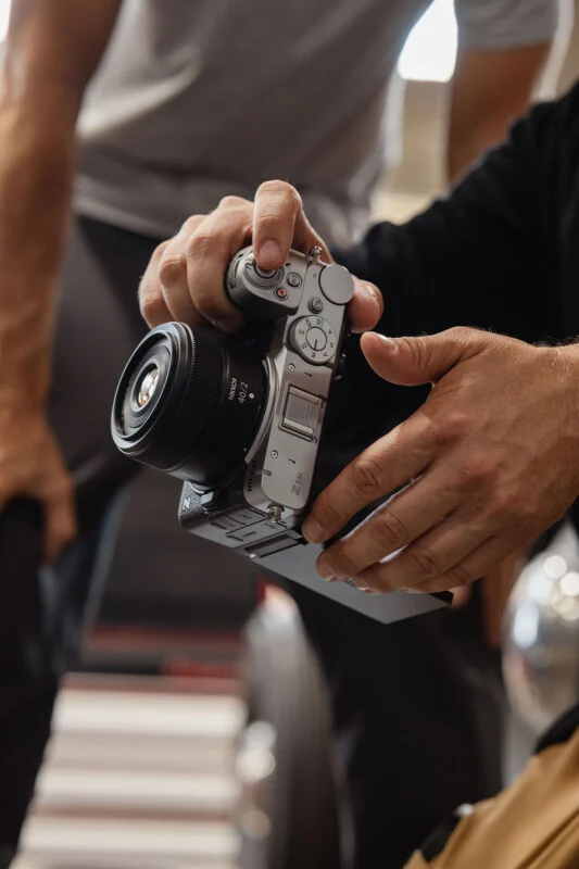A person holds a silver Nikon Z fc camera with a Nikkor 40/2 lens, with another person visible in the blurred background.