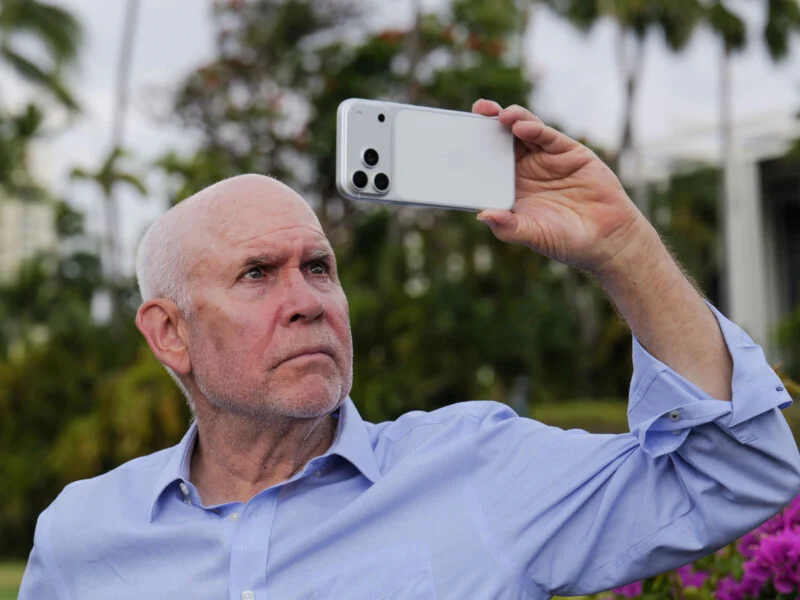 An older man with a shaved head and light blue button-down shirt holds a white smartphone up to take a picture outdoors.