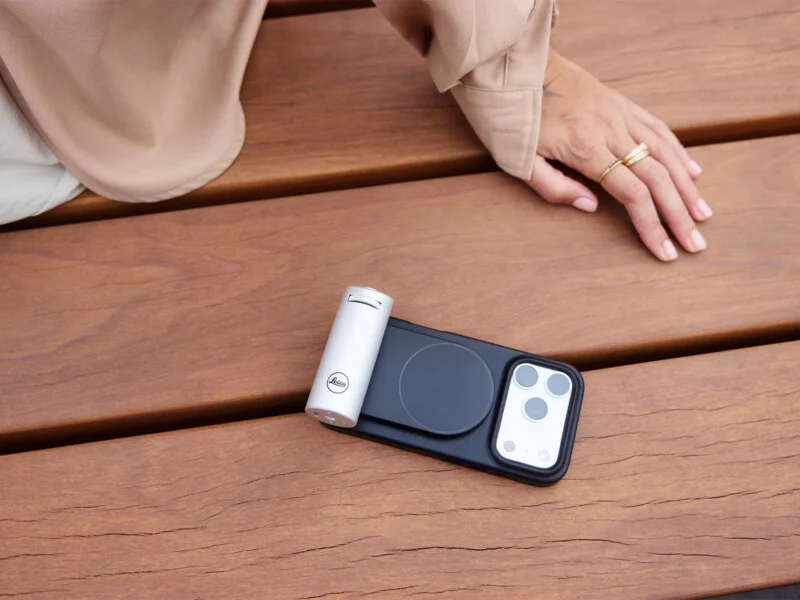A person's hand rests on a wooden bench next to a smartphone in a black case with a silver Leica-branded camera grip attached to the back.
