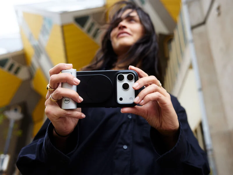 A person holds a smartphone equipped with a camera grip attachment, featuring a visible Leica logo, while standing in front of yellow cube-shaped buildings.