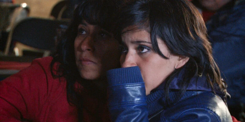 Two women with dark hair sit side-by-side, looking intently toward the right, with one woman wearing a red jacket and the other a blue leather jacket.