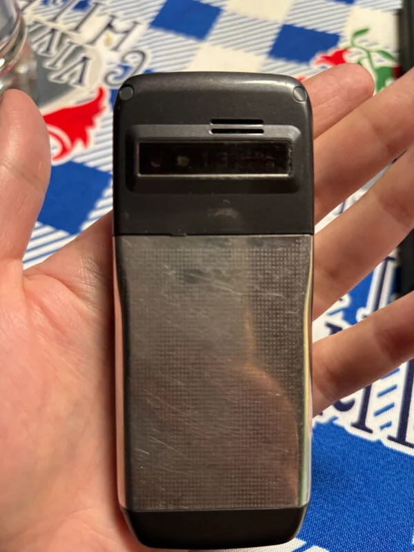A hand holds a rectangular, silver and black mobile phone with a 1.3-megapixel camera lens visible on the back.