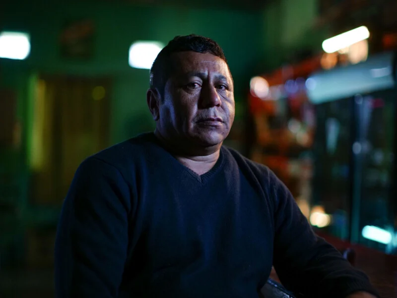 A middle-aged man with short dark hair and a dark V-neck sweater sits in a dimly lit room with green walls.