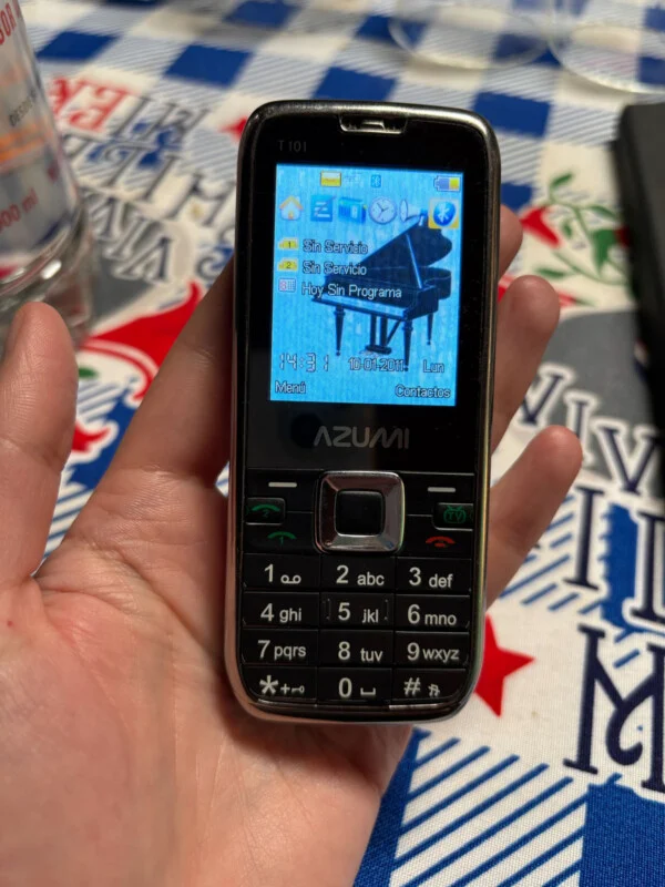 A person holds an Azumi T101 feature phone displaying a piano wallpaper, a date of January 10, 2011, and no service status.