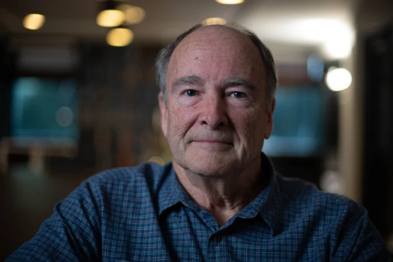 A middle-aged man with thinning hair and a blue plaid shirt looks directly at the camera in a dimly lit room.