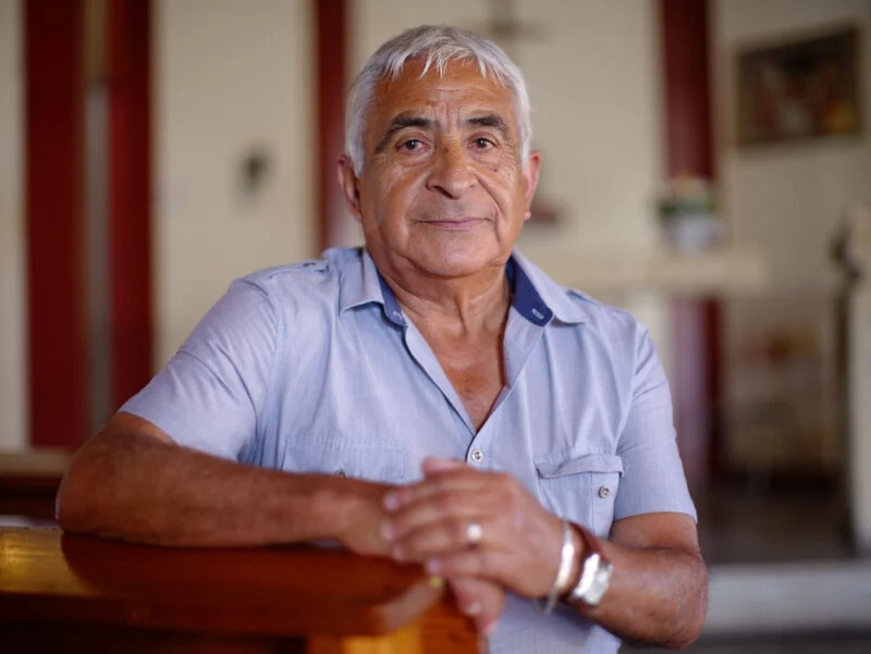 An elderly man with short white hair and a light blue button-down shirt sits with his hands clasped, resting on a wooden pew inside a church.