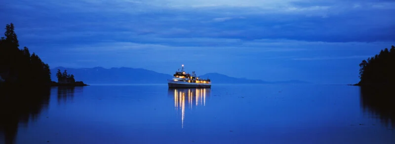 A boat with lights reflecting on calm blue water is anchored between tree-lined shores at dusk, with distant mountains under a cloudy sky.