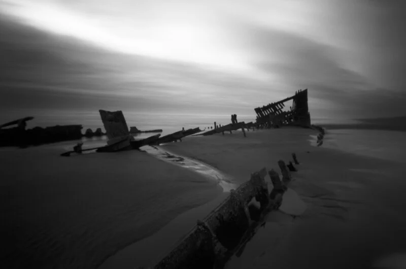 A black and white long-exposure shot of a shipwreck skeleton resting on a sandy beach under a cloudy sky.