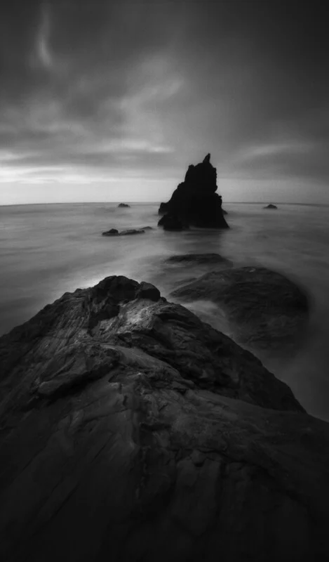 A black and white long-exposure shot of a jagged rock formation rising from a calm, misty ocean with large rocks in the foreground.