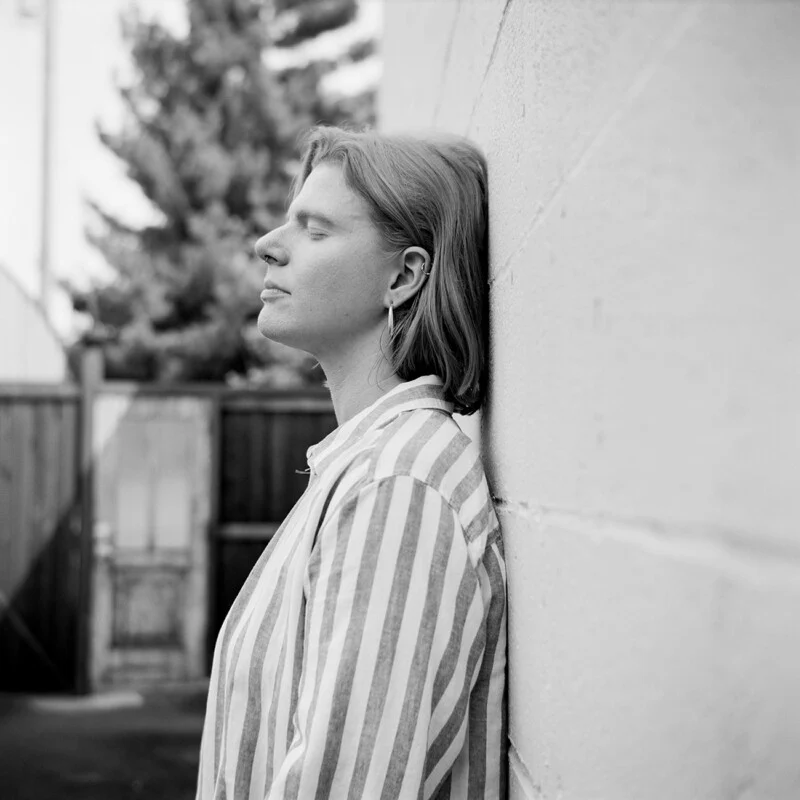 A person with short hair and closed eyes leans against a white brick wall, wearing a striped button-down shirt.