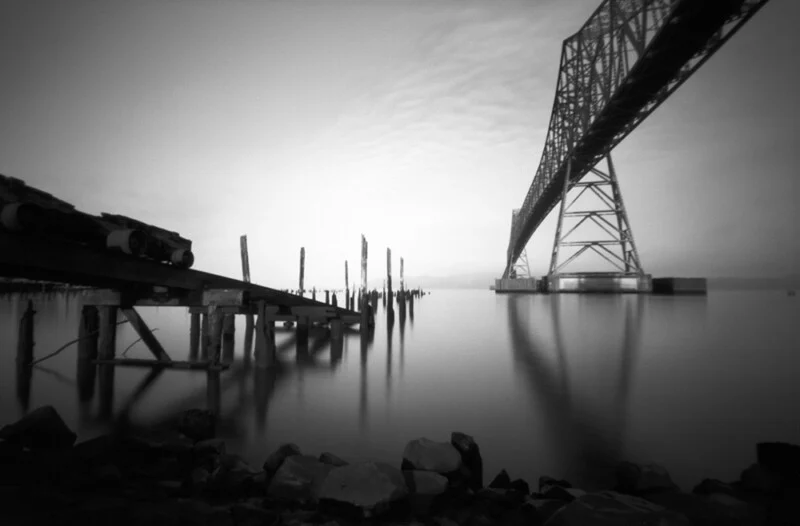 A black-and-white long exposure shot shows a calm body of water with a large steel truss bridge on the right and the remains of a wooden pier on the left.