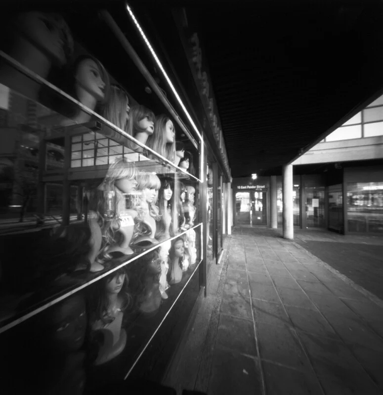 A black and white street-level view of a storefront window displaying rows of mannequin heads wearing wigs, with a sidewalk and building entrance visible to the right.