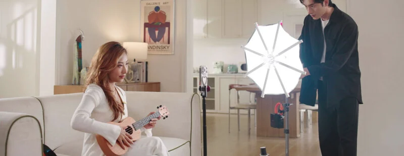 A woman in white sits on a sofa playing a ukulele while a man stands nearby holding a large, illuminated photography umbrella light.