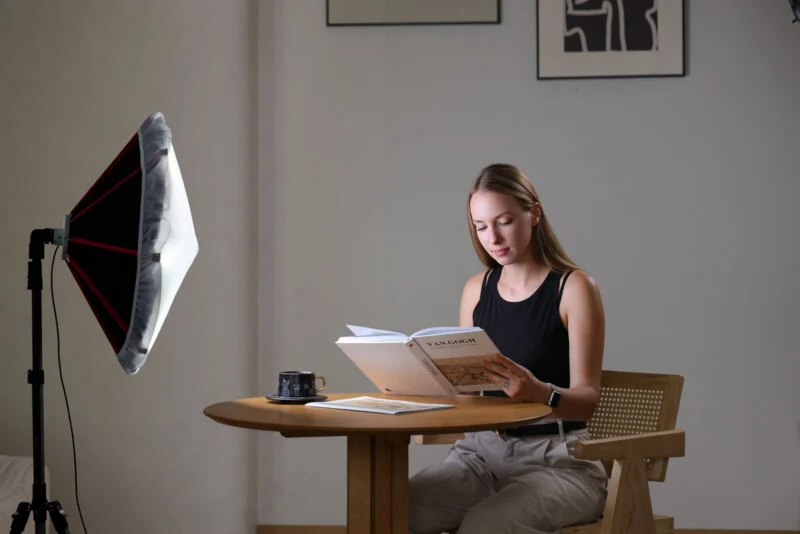 A woman sits at a round wooden table reading a Van Gogh book, illuminated by a large studio softbox light on a stand.