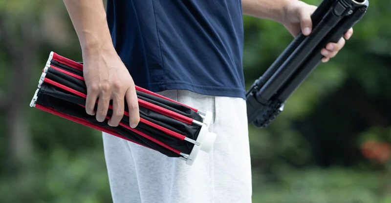 A person in a blue t-shirt and grey sweatpants holds a folded black and red photography softbox in one hand and a tripod in the other.
