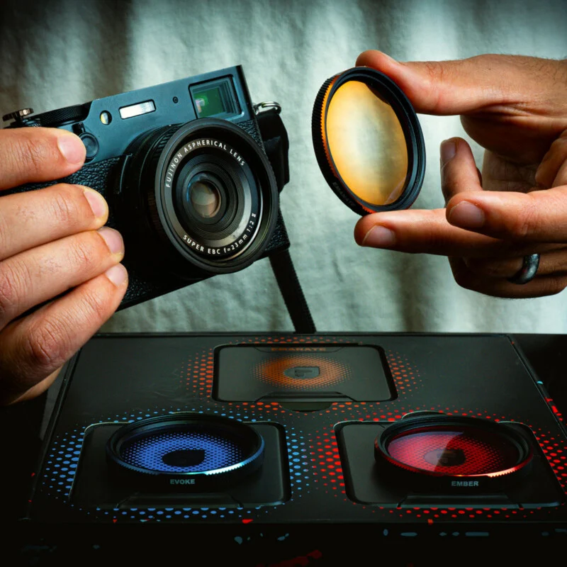 A person holds a black camera in one hand and a circular amber lens filter in the other, with a box containing blue and red filters resting on a surface below.